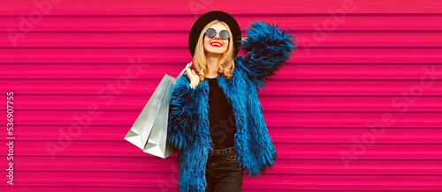 Portrait of beautiful stylish smiling woman with shopping bags wearing blue fur coat, black round hat and sunglasses posing on pink background © rohappy