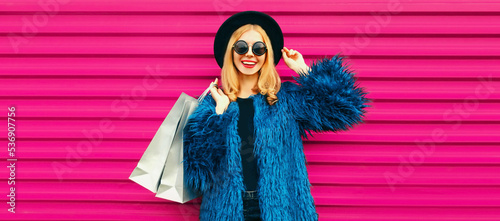Portrait of beautiful stylish smiling woman with shopping bags wearing blue fur coat, black round hat and sunglasses posing on pink background © rohappy