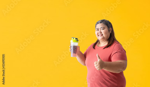 Fat woman holding a water or supplement bottle and thumbs up, isolated on yellow background. Clipping paths for design work empty free space © kromkrathog