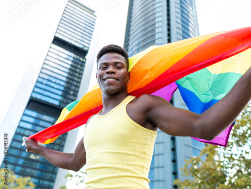 Smiling young man wearing yellow vest holding rainbow flag in front of modern buildings photo