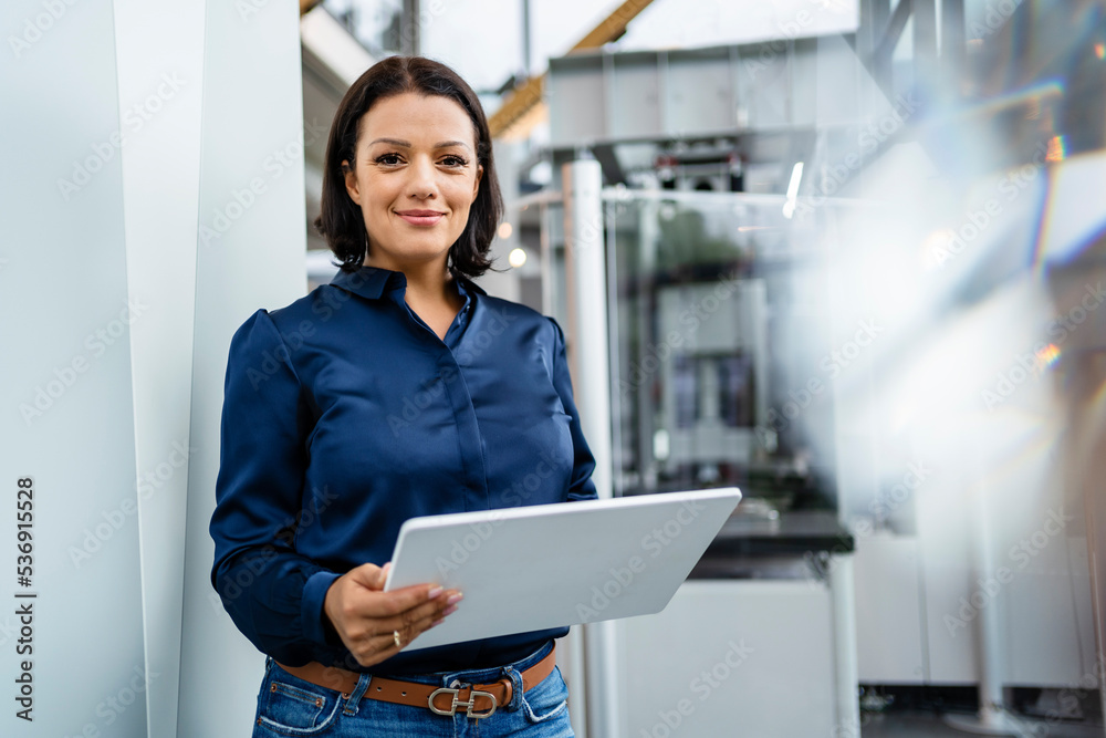 Smiling businesswoman holding tablet PC standing by wall in industry