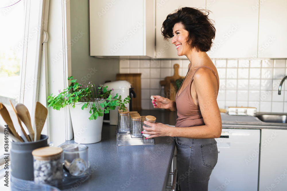 Smiling woman holding jar looking through window standing at kitchen counter Smiling woman holding jar looking through window standing at kitchen counter