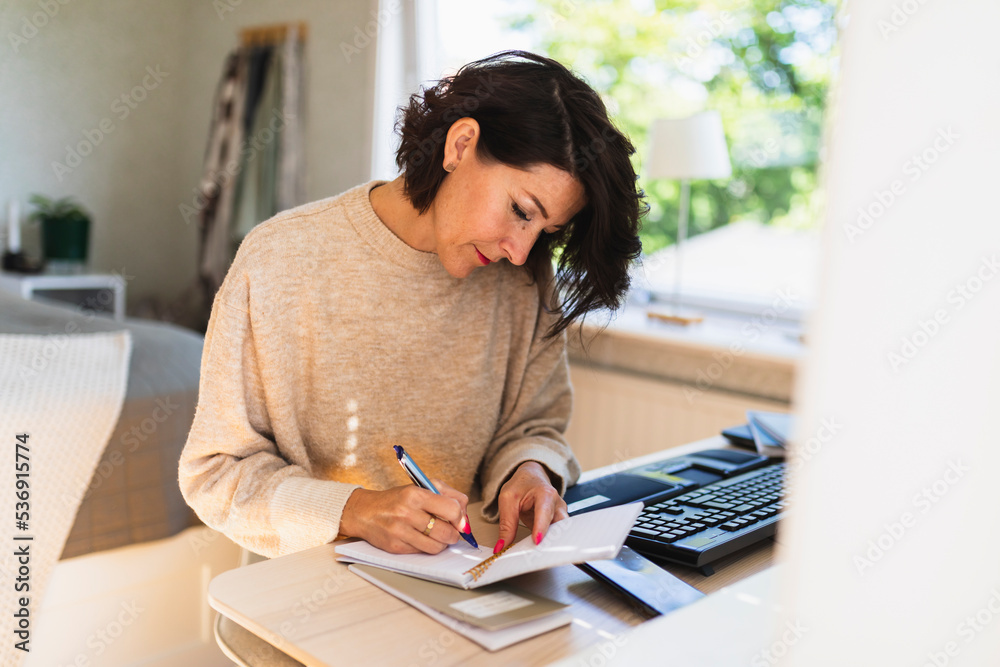 Mature businesswoman writing in note pad on table at home Mature businesswoman writing in note pad on table at home