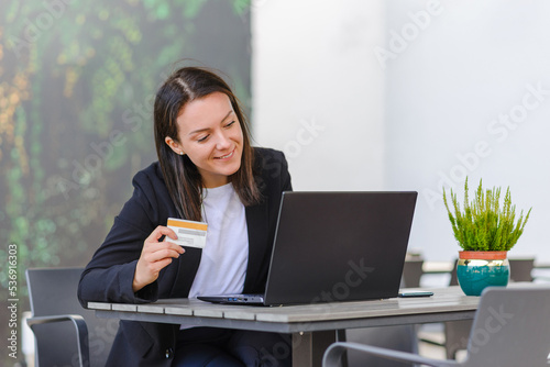 Young smiling business woman holding in hand credit bank card and paying online while sitting at cafe table with laptop, sitisfied female office employee shopping buying goods in internet. photo