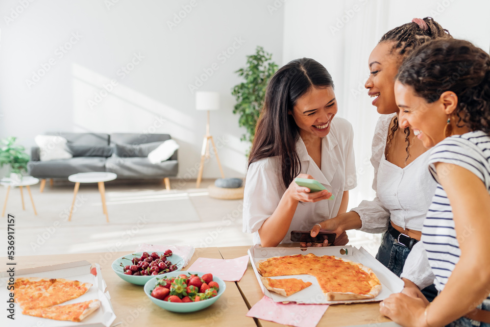 Cheerful roommates having pizza and using smart phone at home