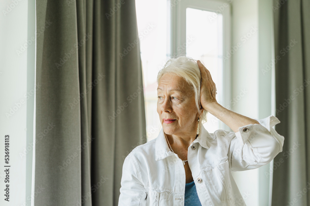 Senior woman touching white hair near curtain at home Senior woman touching white hair near curtain at home