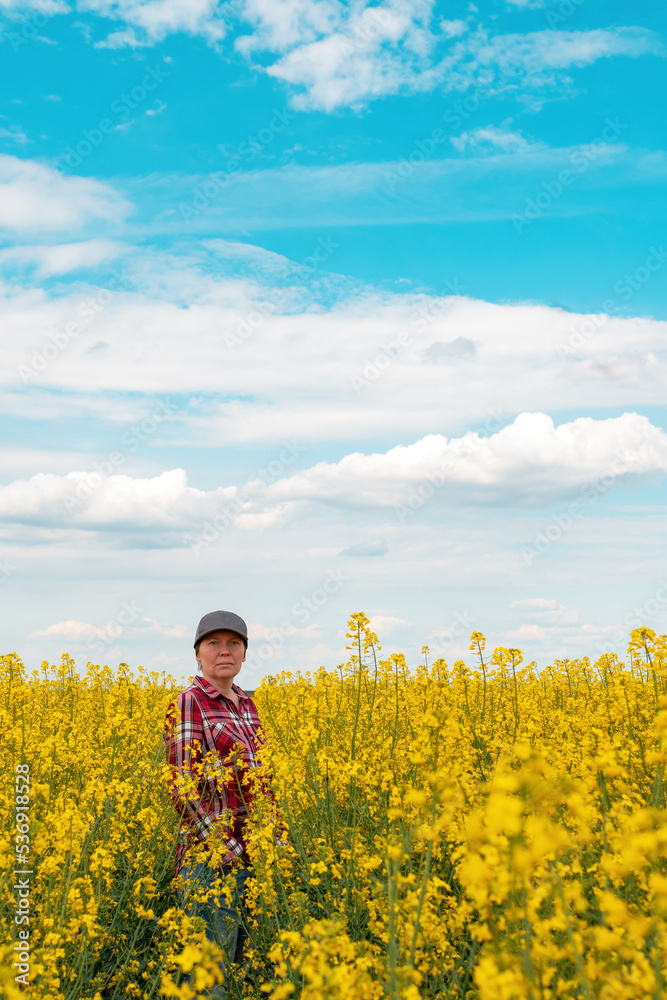 Female farm worker wearing red plaid shirt and trucker's hat standing in cultivated rapeseed field in bloom and looking over crops