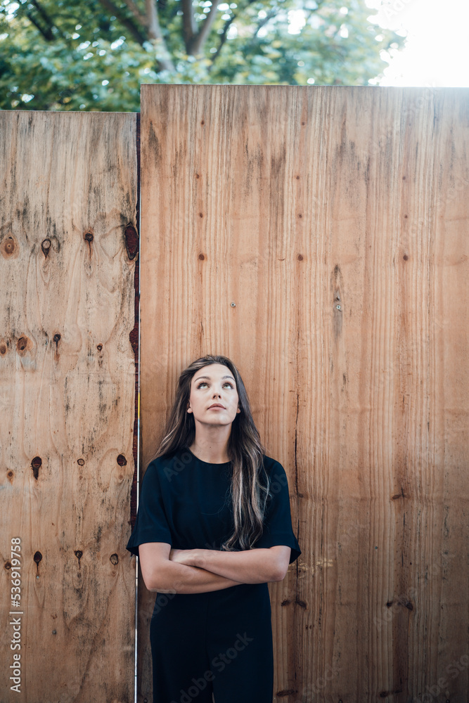 Young woman standing with arms crossed looking up in front of wooden wall Young woman standing with arms crossed looking up in front of wooden wall