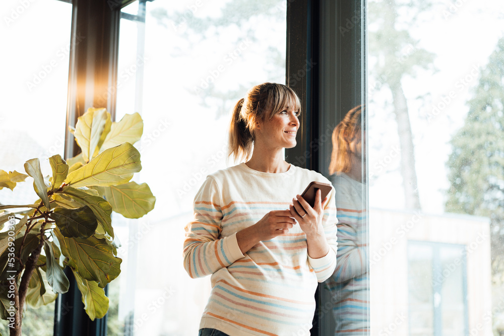 © Westend61 - Smiling pregnant woman holding mobile phone leaning on glass door © Westend61 - Smiling pregnant woman holding mobile phone leaning on glass door