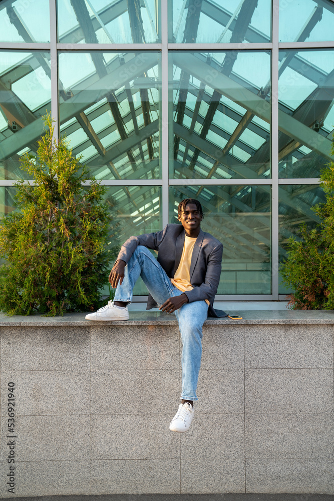 Young businessman sitting on wall in front of glass house Young businessman sitting on wall in front of glass house