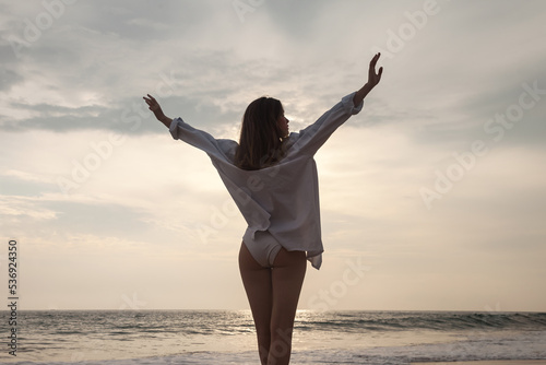 Rear view happy young woman in casual white shirt arms raised at ocean sandy beach. Relaxing, fun and enjoy holiday at tropical seashore. Concept of beach holidays, summer vacation. Copy text space