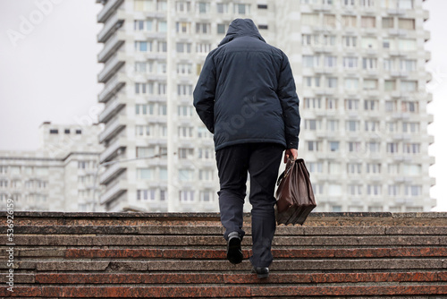 Foto Man in hooded jacket with leather briefcase climbing stone stairs in city during rain