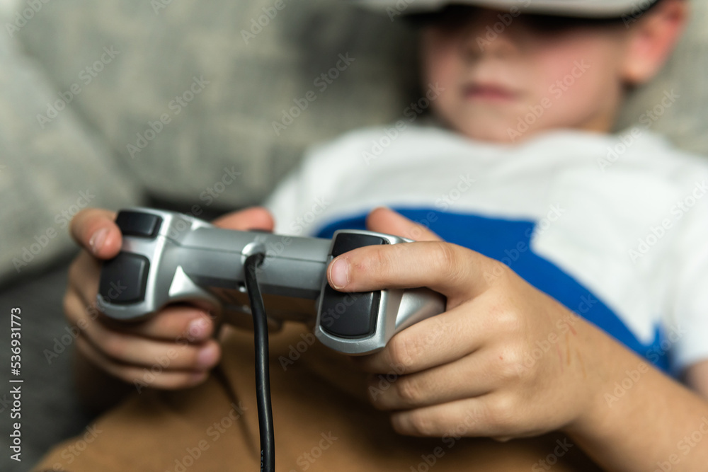 Child lying on the sofa with virtual reality goggles connected to a game console controller playing an online game. Child lying on the sofa with virtual reality goggles connected to a game console controller playing an online game.