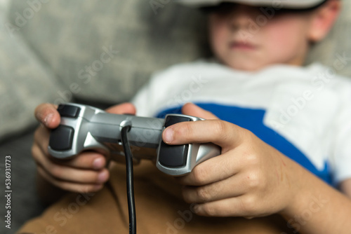 Child lying on the sofa with virtual reality goggles connected to a game console controller playing an online game. © Jorge