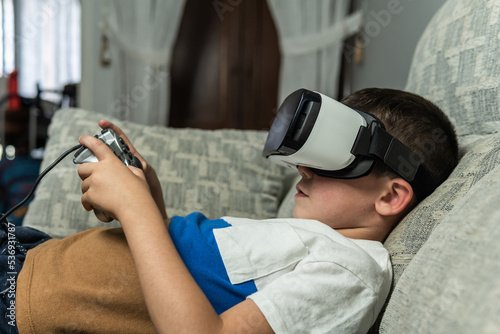 Side image of a child lying on the sofa with virtual reality goggles connected to a game console controller playing an online game.