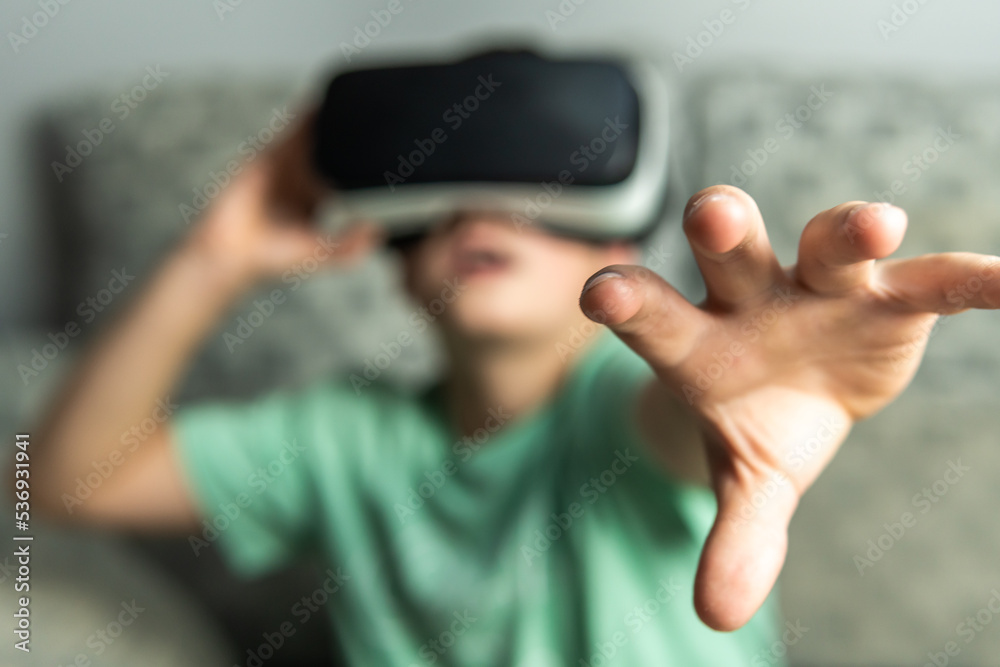 Front shot of a dark-haired boy holding his virtual reality goggles and putting a hand in front to touch the generated 3D space.