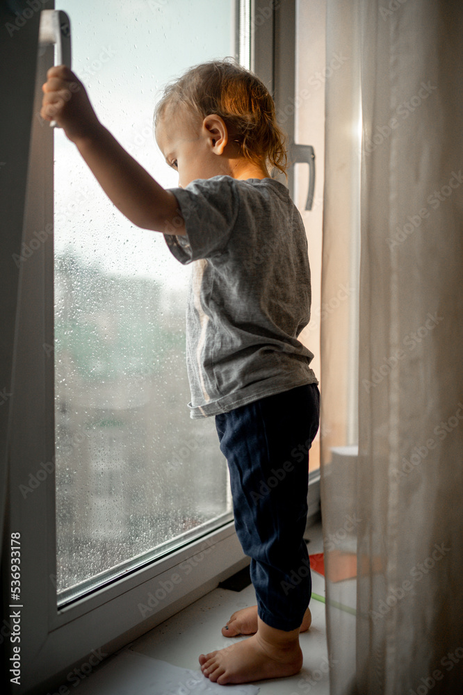 The baby is standing at the window on the windowsill. Curious little boy looks out the window, standing at home on the windowsill