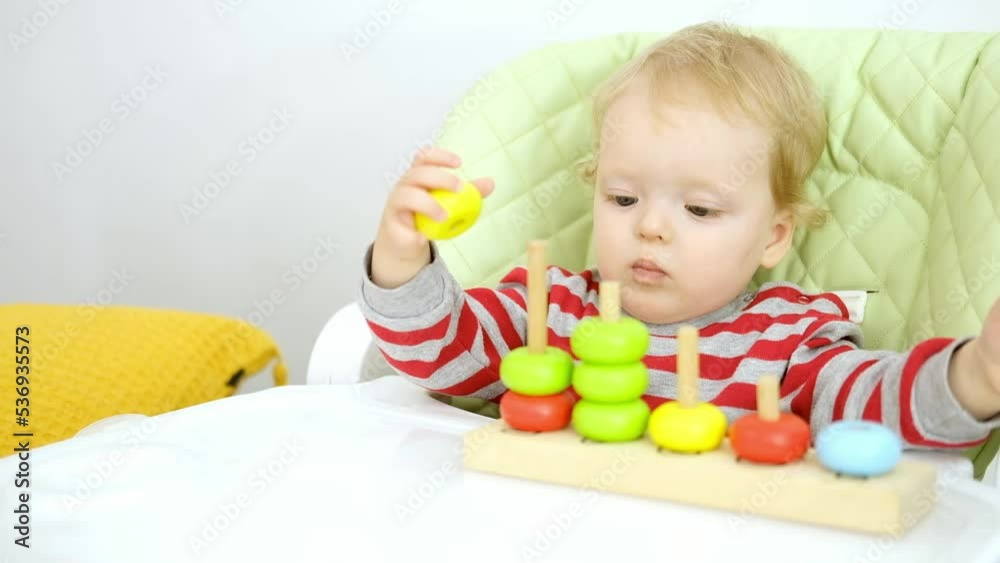 Funny serious cute Caucasian baby girl is sitting on high chair and playing with educational wooden toy. Concept of early learning and development of fine motor skills. Funny serious cute Caucasian baby girl is sitting on high chair and playing with educational wooden toy. Concept of early learning and development of fine motor skills.