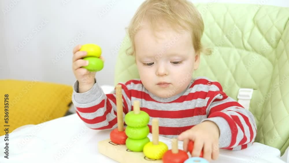 Funny serious cute Caucasian baby girl is sitting on high chair and playing with educational wooden toy. Concept of early learning and development of fine motor skills. Funny serious cute Caucasian baby girl is sitting on high chair and playing with educational wooden toy. Concept of early learning and development of fine motor skills.
