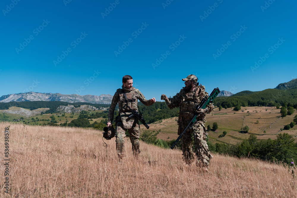 A sniper team squad of soldiers is going undercover. Sniper assistant and team leader walking and aiming in nature with yellow grass and blue sky. Tactical camouflage uniform. A sniper team squad of soldiers is going undercover. Sniper assistant and team leader walking and aiming in nature with yellow grass and blue sky. Tactical camouflage uniform.
