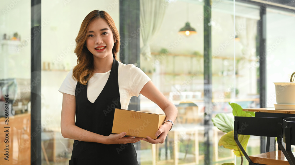 Fototapeta premium Beautiful female entrepreneur wearing apron holding cardboard and smiling to camera. Online selling, E-commerce concept