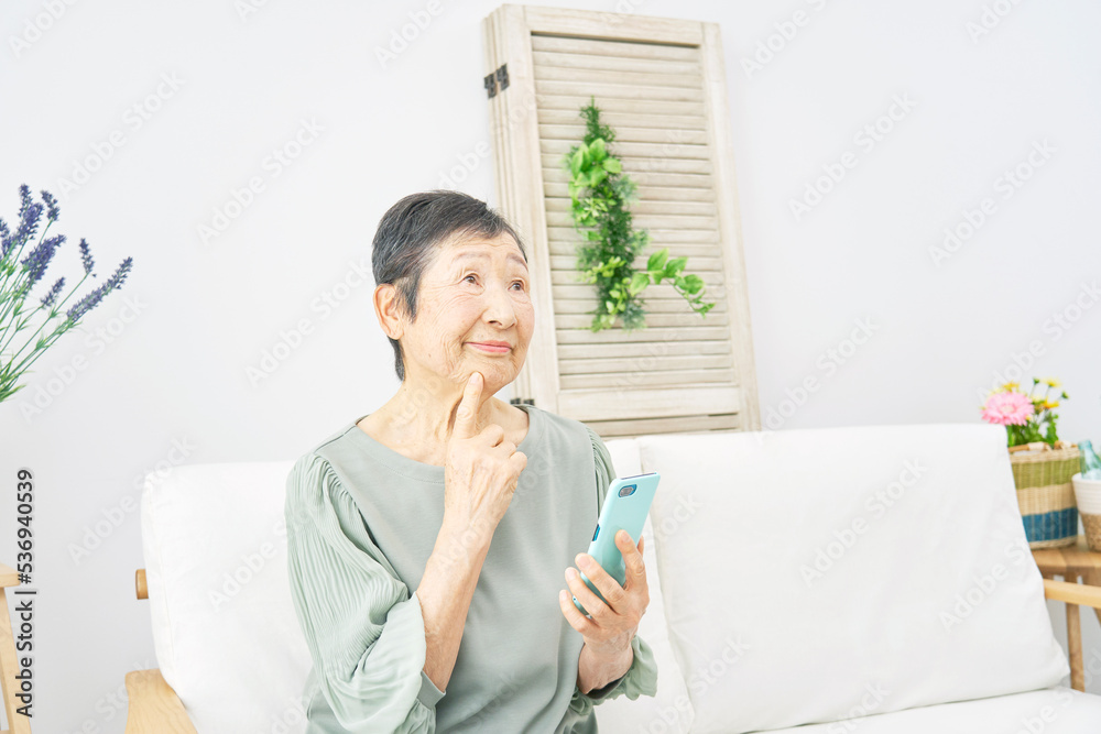 リビングでスマホを持って考える高齢者女性 リビングでスマホを持って考える高齢者女性