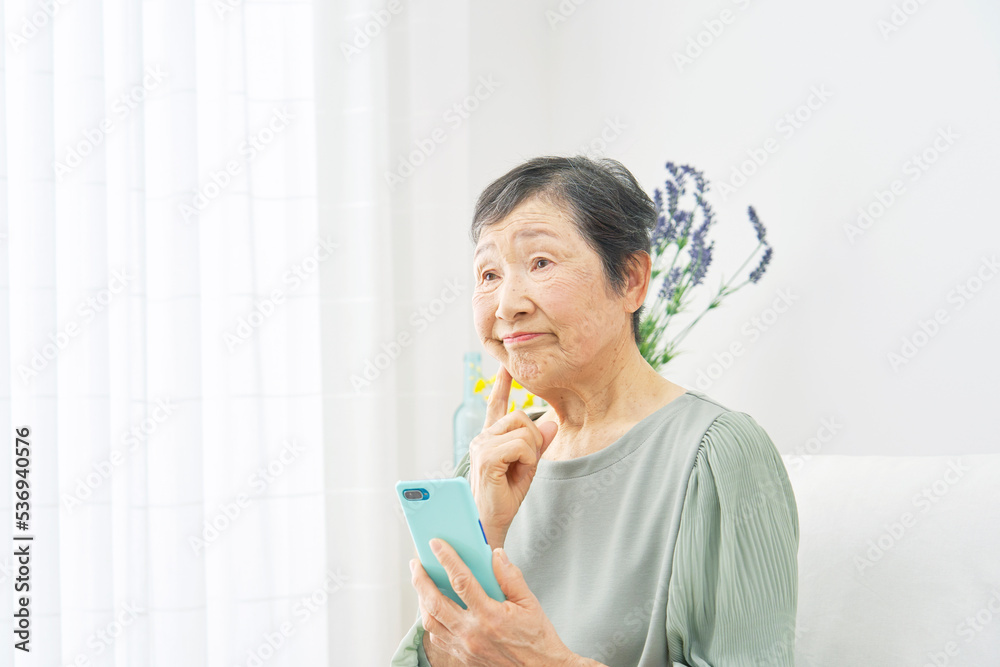 リビングでスマホを持って考える高齢者女性 リビングでスマホを持って考える高齢者女性