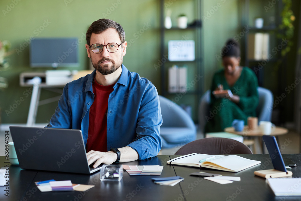 Young serious businessman in eyeglasses looking at camera while networking or preparing presentation by workplace in front of laptop