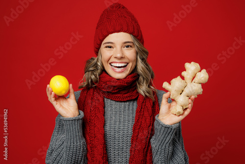 Young smiling happy fun woman wear grey sweater scarf hat hold show lemon ginger root isolated on plain red background studio portrait Healthy lifestyle ill sick disease treatment cold season concept photo