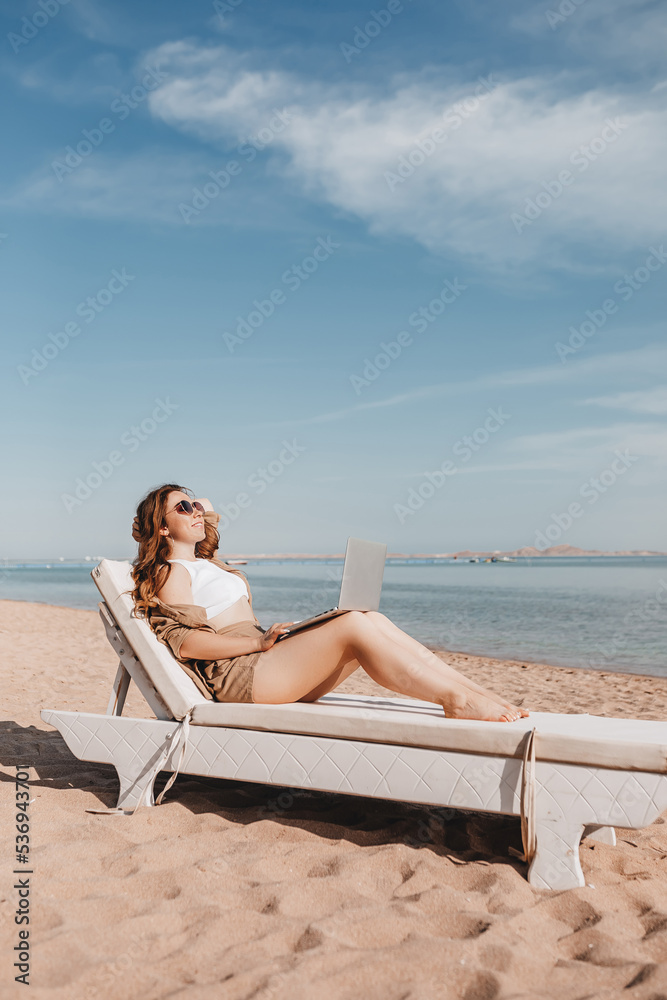 A cheerful blogger girl remotely works with a laptop on the beach with sea views. A woman makes a video from a trip. Travels the world and works with social networks online. Remote modern profession