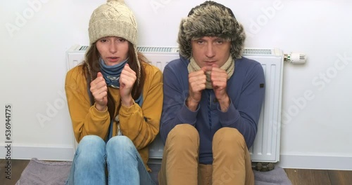 Funny couple man, woman shaking from cold at white battery heater background. Young people freezing in home room, dressed in warm clothes. Heating apartments, houses problems. Gas, electricity lack