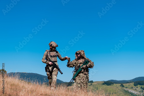 A sniper team squad of soldiers is going undercover. Sniper assistant and team leader walking and aiming in nature with yellow grass and blue sky. Tactical camouflage uniform.