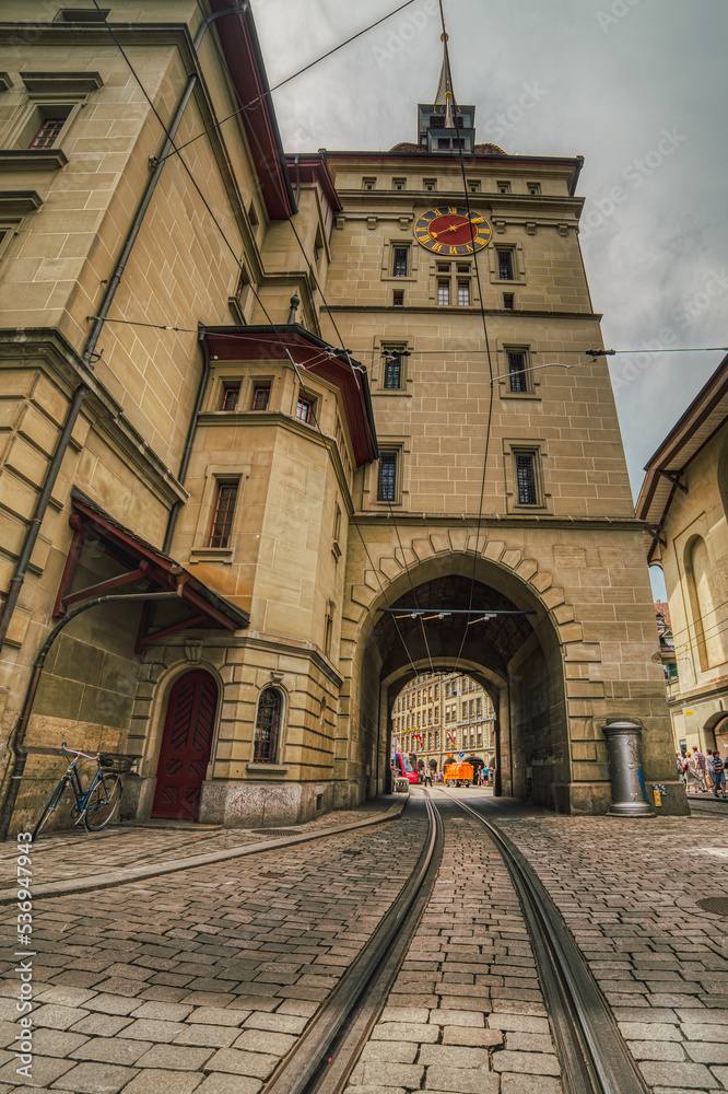 Tram railway tunnel under the baroque tower Kafigturm above a old historical cobbled street. Cablecar track or tracks across the arched medieval steeple in the popular Marktgasse. Bern, Switzerland