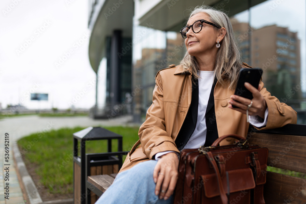 a stylish elderly woman with a mobile phone in her hands rests on a bench and looks around