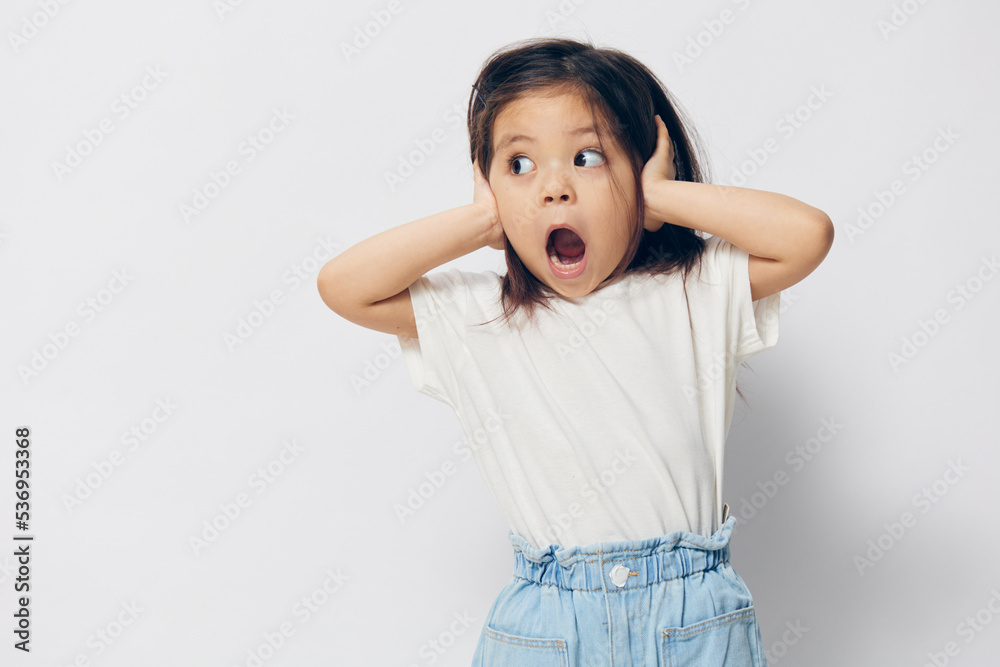 a cute little girl of preschool age in a white T-shirt, standing on a white background, screaming, covering her ears with her hands and looking away in fright. The theme of children's fright a cute little girl of preschool age in a white T-shirt, standing on a white background, screaming, covering her ears with her hands and looking away in fright. The theme of children's fright