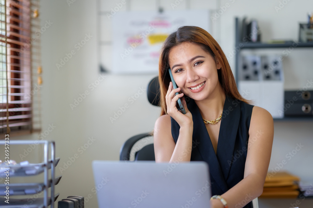 Asian businesswoman sitting with her laptop happily chatting on the phone with a customer explaining in detail the company's products in the office. Asian businesswoman sitting with her laptop happily chatting on the phone with a customer explaining in detail the company's products in the office.