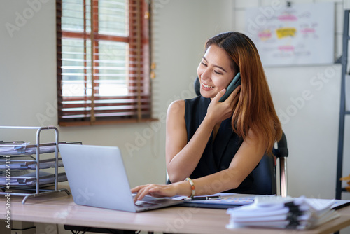 Asian businesswoman sitting with her laptop happily chatting on the phone with a customer explaining in detail the company's products in the office.