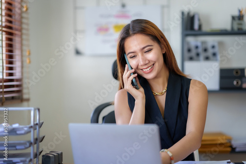 Asian businesswoman sitting with her laptop happily chatting on the phone with a customer explaining in detail the company's products in the office.