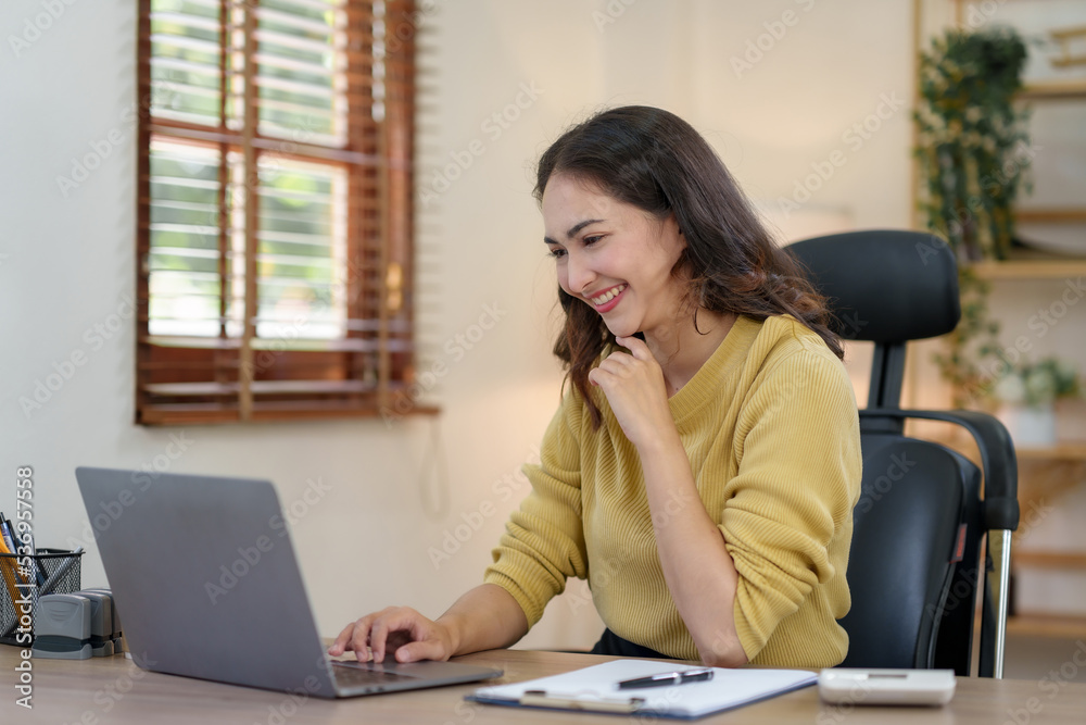 Beautiful young Asian female sitting using a laptop at home. Happy business woman smiling and enjoying her work and taking note on a notepad.