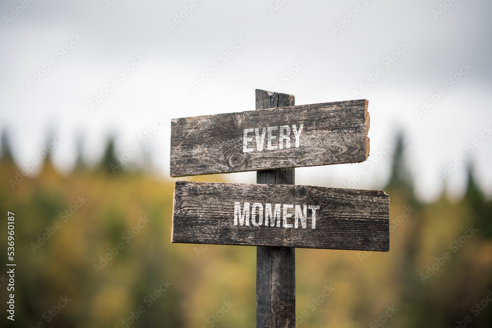 vintage and rustic wooden signpost with the weathered text quote every moment, outdoors in nature. blurred out forest fall colors in the background.