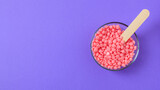 Pink granules of wax for depilation in a glass bowl on a lilac background. Place for text. Epilation, depilation, waxing, unwanted hair removal procedure.