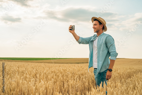 Happy farmer photographing crops with phone while standing in his growing wheat field. 