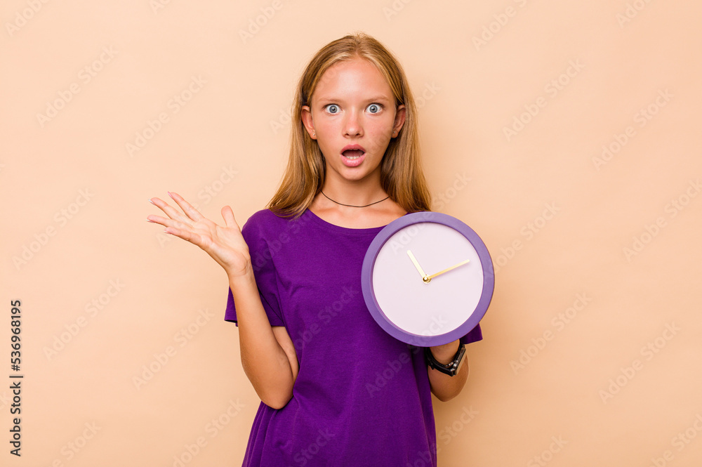 Little caucasian girl holding a clock isolated on beige background surprised and shocked. Little caucasian girl holding a clock isolated on beige background surprised and shocked.