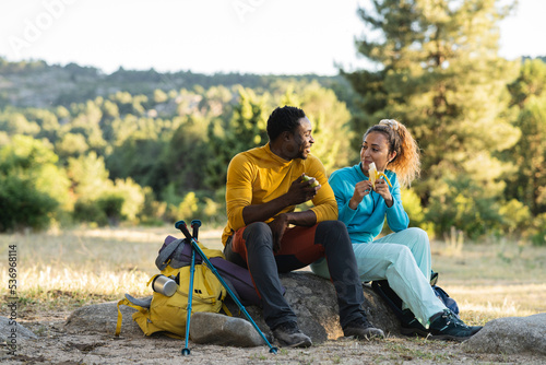 couple in nature having lunch after hiking. they are eating fruit sitting on a rock at sunset while talking.