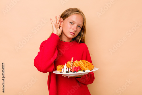 Little caucasian girl holding a waffles isolated on beige background trying to listening a gossip.
