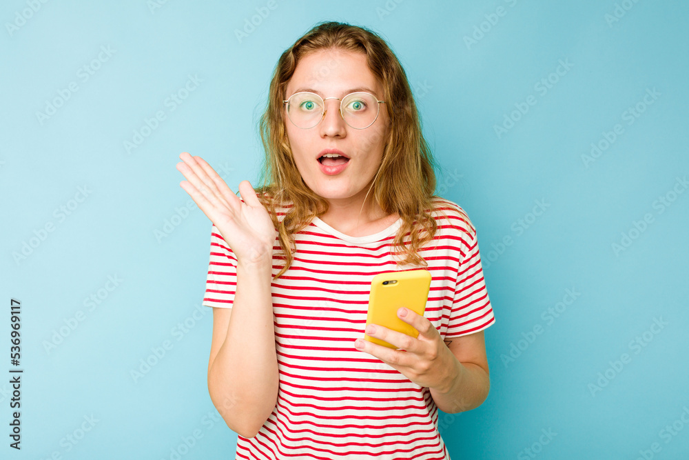 Young caucasian woman holding mobile phone isolated on blue background surprised and shocked. Young caucasian woman holding mobile phone isolated on blue background surprised and shocked.