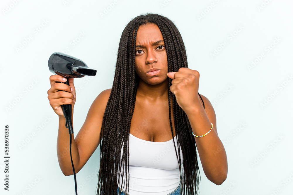Young African American woman holding a hairdryer isolated on blue background showing fist to camera, aggressive facial expression. Young African American woman holding a hairdryer isolated on blue background showing fist to camera, aggressive facial expression.