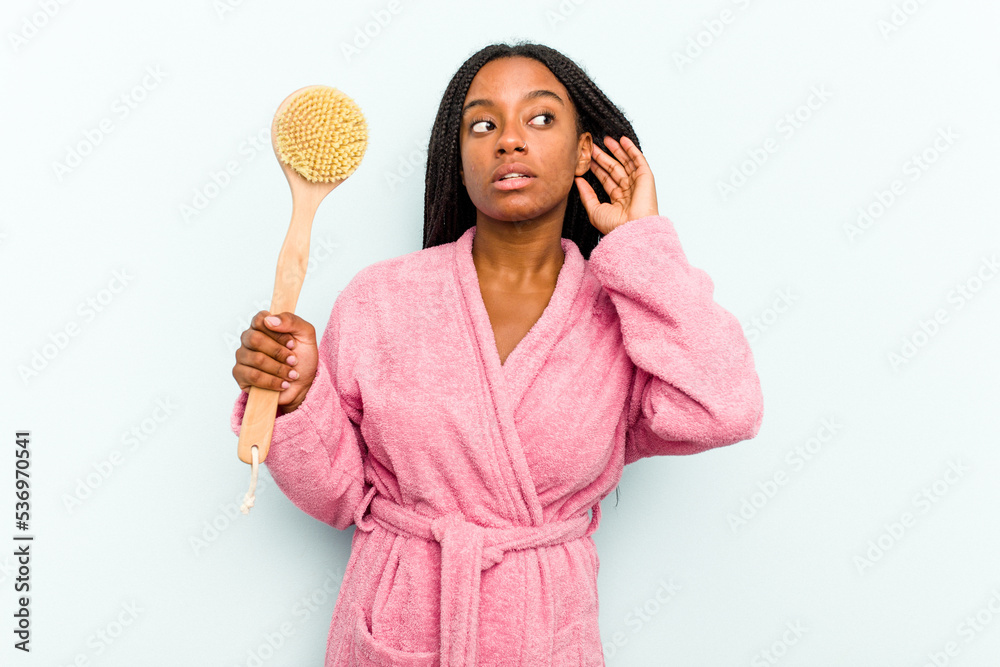 Young African American woman holding a bathtub brush isolated on blue background trying to listening a gossip. Young African American woman holding a bathtub brush isolated on blue background trying to listening a gossip.