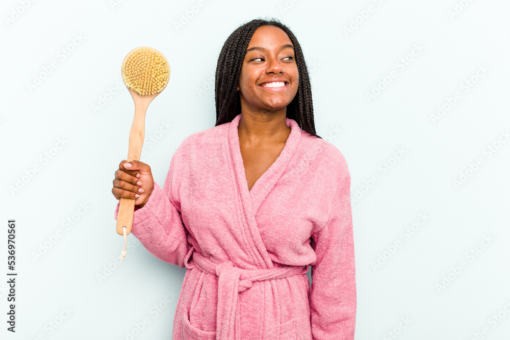 Young African American woman holding a bathtub brush isolated on blue background looks aside smiling, cheerful and pleasant. Young African American woman holding a bathtub brush isolated on blue background looks aside smiling, cheerful and pleasant.