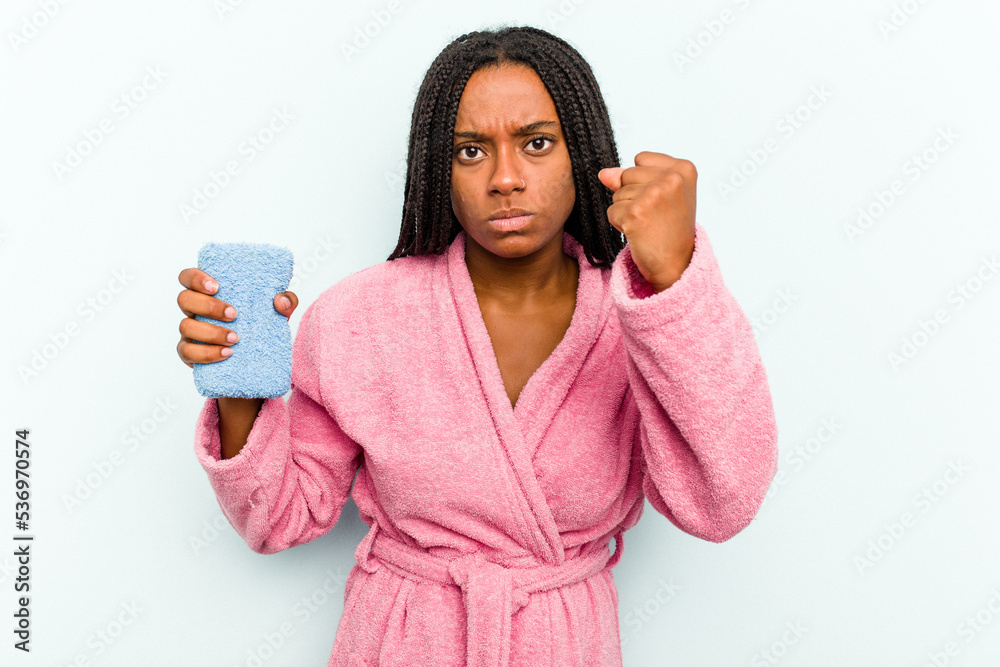 Young African American woman holding a sponge isolated on blue background showing fist to camera, aggressive facial expression. Young African American woman holding a sponge isolated on blue background showing fist to camera, aggressive facial expression.